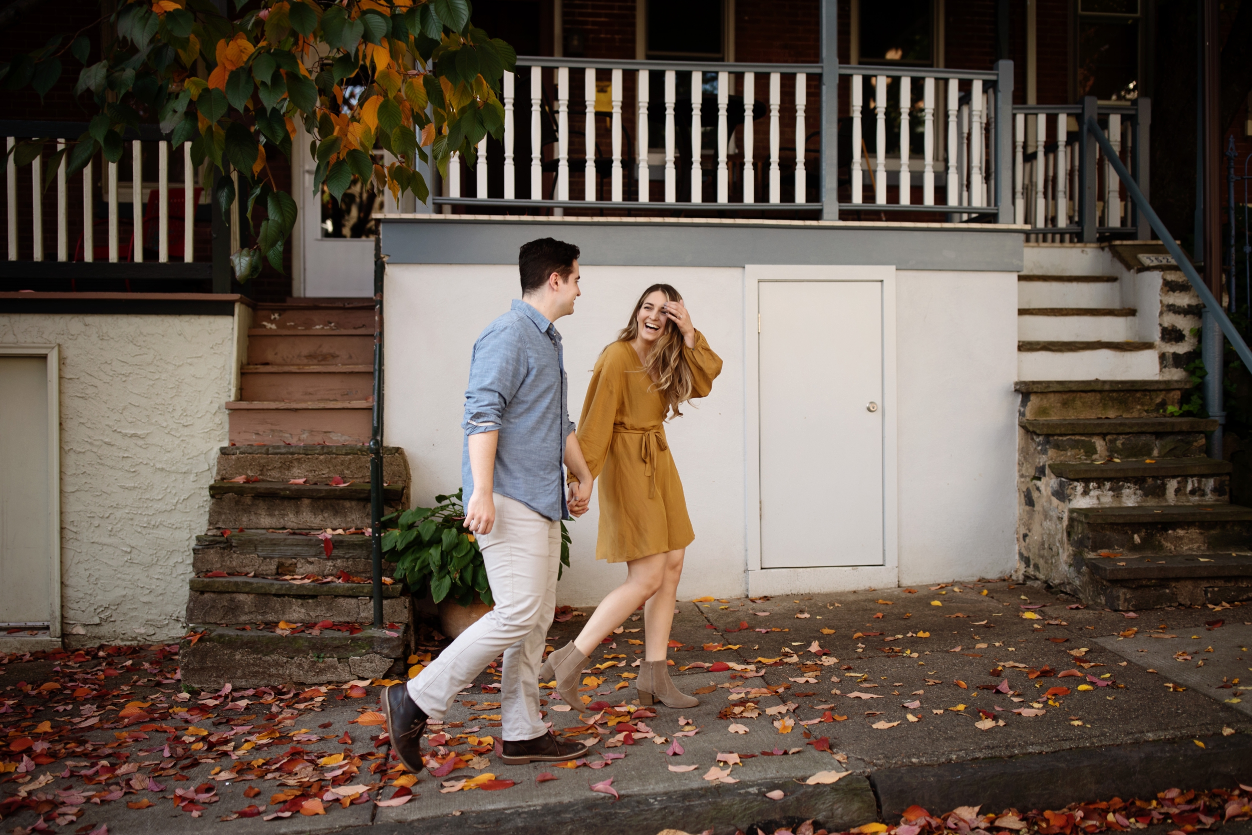 East Falls Philadelphia Engagement Photo Session Couple strolling through their neighbor in very authentic and romantic fall engagement photos. A woman wearing a wrapped mustard yellow dress and the man wearing denim blue button up and khaki pants, strolling through their Philadelphia neighborhood with their puppy.