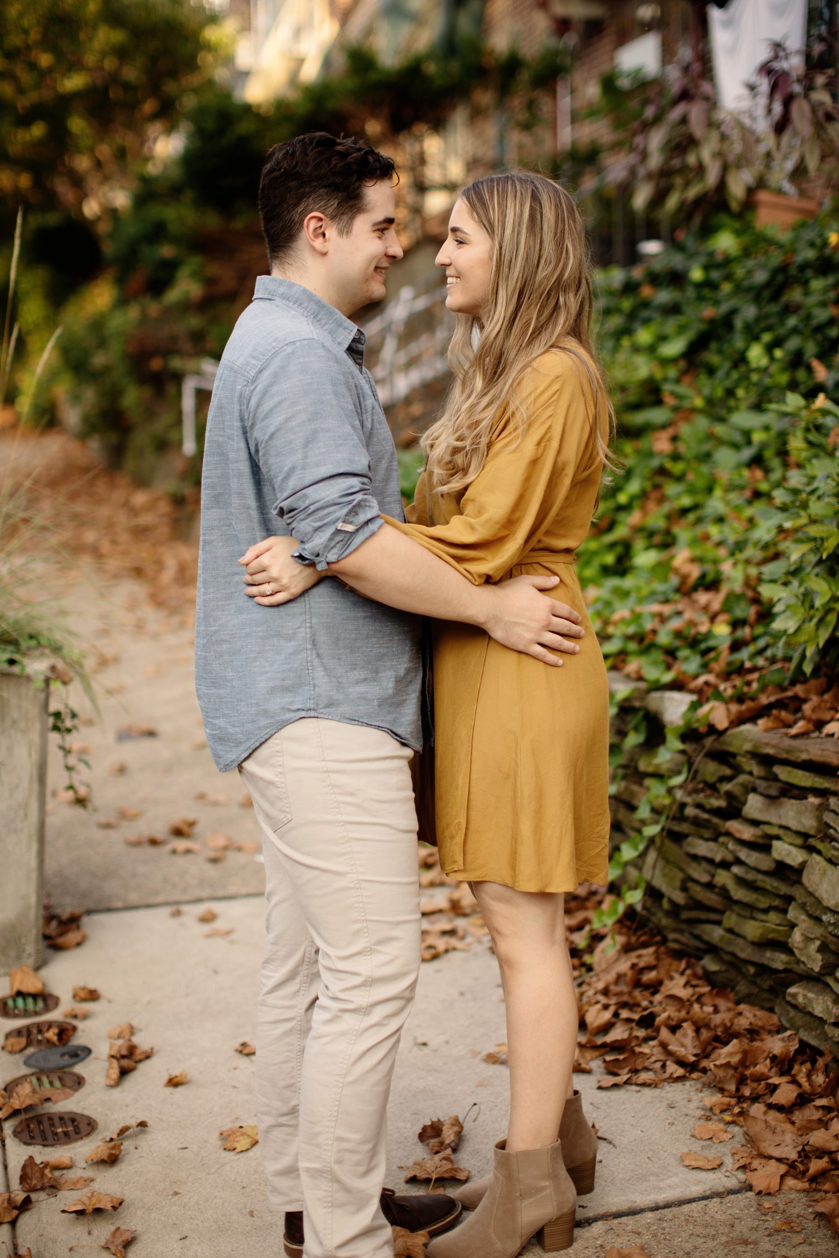 East Falls Philadelphia Engagement Photo Session Couple strolling through their neighbor in very authentic and romantic fall engagement photos. A woman wearing a wrapped mustard yellow dress and the man wearing denim blue button up and khaki pants, strolling through their Philadelphia neighborhood with their puppy.